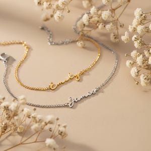 Puede incluir: Se muestran dos delicadas pulseras con nombres, una dorada y otra plateada. La pulsera dorada deletrea "emily" en cursiva, mientras que la pulsera plateada deletrea "james". Las pulseras están sobre un fondo neutro con flores blancas.