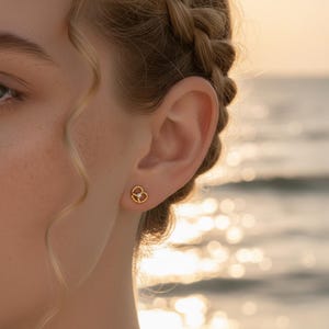 May include: Gold-coloured stud earrings in a pretzel design, each with a small, clear gemstone at the centre. The earrings are shown on a person's ear, with a blurred, sunlit background.