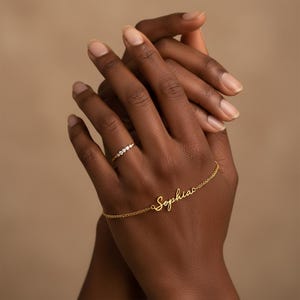 May include: A gold-colored ring with three small, clear stones and a gold bracelet with the name "Sophia" in cursive script. The jewelry is displayed on a hand with a neutral background.