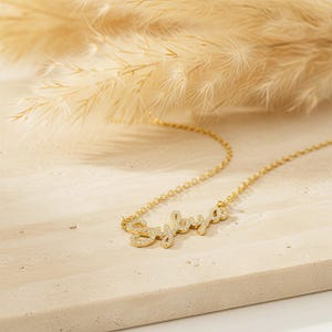 May include: A gold necklace featuring the name "Sylvia" in a cursive font, with the letters embellished with small, clear gemstones. The necklace is displayed on a light-toned, textured surface, with dried ornamental grass in the background.
