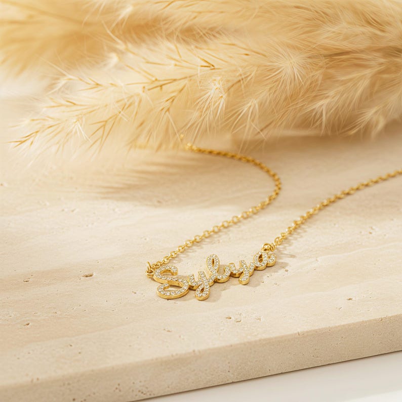May include: A gold necklace featuring the name "Sylvia" in a cursive font, with the letters embellished with small, clear gemstones. The necklace is displayed on a light-toned, textured surface, with dried ornamental grass in the background.