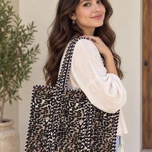 May include: A rectangular tote bag featuring a brown and black leopard print pattern. The bag has structured sides and is accented with black and white striped fabric trim along the edges and the two shoulder straps, providing a distinct contrast.