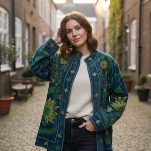 May include: A teal jacket with green and yellow floral embroidery. The jacket is worn over a cream sweater and black jeans. The design includes a stand-up collar and no buttons. The jacket's length is mid-hip.