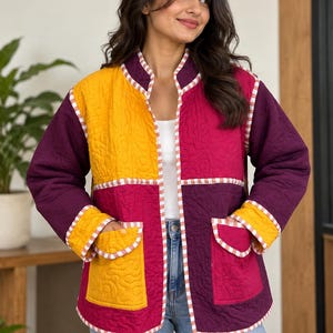 Può includere: Una giacca trapuntata con un design a blocchi di colore in giallo senape, magenta e viola scuro. Il capo presenta un colletto alla coreana, maniche lunghe, due tasche frontali applicate e bordi rifiniti con un profilo a scacchi bianchi e rossi.