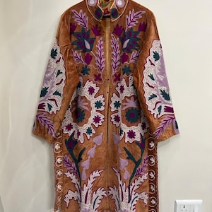 May include: A long, tan-colored coat featuring intricate, multi-colored embroidery in shades of deep purple, dark green, and white. The garment has a high collar, long sleeves, and a front-open design, showcasing floral and leaf patterns.