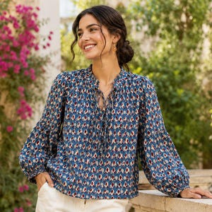 May include: A long-sleeved blouse in a medium blue hue featuring a repeating diamond-shaped pattern in shades of dark red, white, and black. The garment includes a gathered neckline with a central tie closure and loose, billowy sleeves.