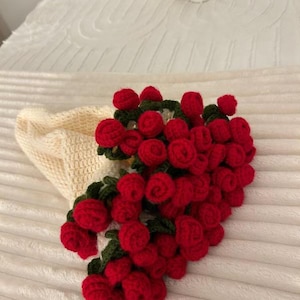 May include: A handmade crochet bouquet of red roses with green leaves, on a cream crocheted base. The roses are densely packed, creating a full, textured appearance. The bouquet is set against a soft, white background.