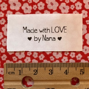Puede incluir: Etiqueta de tela blanca con texto negro que dice "Made with LOVE ❤ by Nana ❤". La etiqueta se mide con una regla, mostrando que tiene aproximadamente 5 cm de ancho.