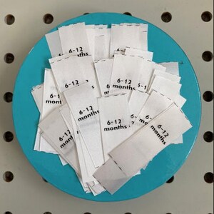 May include: A collection of white fabric labels, each printed with "6-12 months", arranged on a round, turquoise-colored surface. The labels are likely for children's clothing.