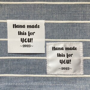 Puede incluir: Dos etiquetas de tela blanca con texto negro. Las etiquetas dicen "Nana made this for you! ~2023~"
