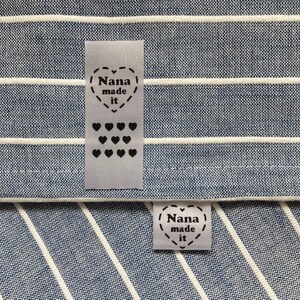 Puede incluir: Dos etiquetas de tela blanca con texto negro y corazones. La etiqueta superior dice "Nana made it" y tiene una fila de 12 corazones peque&ntilde;os. La etiqueta inferior dice "Nana made it" y tiene un coraz&oacute;n alrededor del texto.