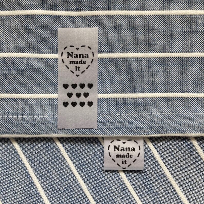 Puede incluir: Dos etiquetas de tela blanca con texto negro y corazones. La etiqueta superior dice "Nana made it" y tiene una fila de 12 corazones peque&ntilde;os. La etiqueta inferior dice "Nana made it" y tiene un coraz&oacute;n alrededor del texto.