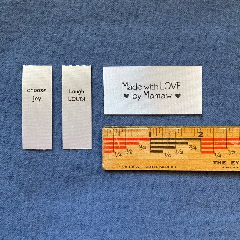 Puede incluir: Tres etiquetas de tela blanca con texto negro. La primera etiqueta dice "choose joy". La segunda etiqueta dice "Laugh LOUD!". La tercera etiqueta dice "Made with LOVE ❤ by Mamaw ❤". Una regla de madera est&aacute; en la esquina inferior derecha de la imagen.