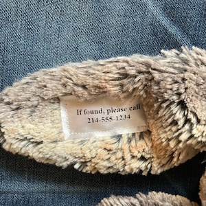 May include: Close-up of a plush toy ear with a tag that reads "If found, please call 214-555-1234." The ear is a mix of brown, beige, and gray faux fur. The background is a blue denim fabric.