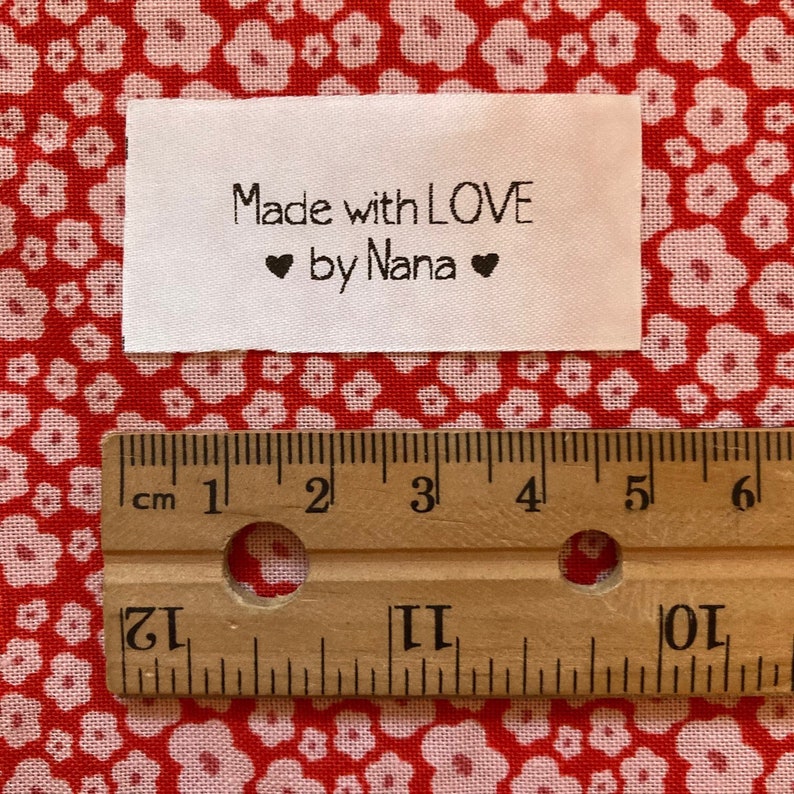 Puede incluir: Etiqueta de tela blanca con texto negro que dice "Made with LOVE ❤ by Nana ❤". La etiqueta se mide con una regla, mostrando que tiene aproximadamente 5 cm de ancho.