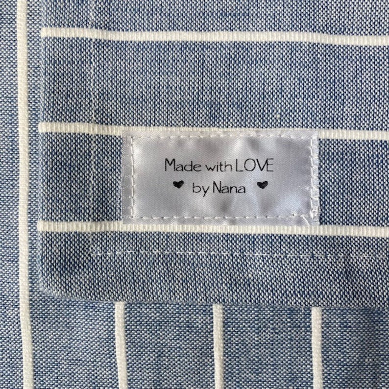 Puede incluir: Una tela a rayas azules y blancas con una etiqueta blanca que dice "Made with LOVE by Nana" con dos peque&ntilde;os corazones a cada lado del texto.
