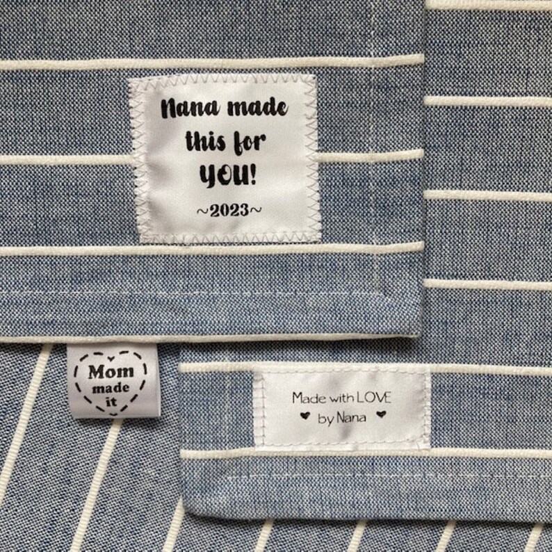 Puede incluir: Dos etiquetas de tela blanca con texto negro sobre una tela a rayas azul y blanca. La etiqueta superior dice "Nana made this for you! ~2023~". La etiqueta inferior dice "Mom made it" y "Made with LOVE by Nana".