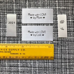 Puede incluir: Cuatro etiquetas de tela blanca con texto negro. Las etiquetas dicen "2021", "Made with LOVE ❤ by Mom ❤", "Made with LOVE ❤ by Lolli ❤" y un s&iacute;mbolo de coraz&oacute;n. Una regla amarilla es visible en la parte inferior de la imagen.