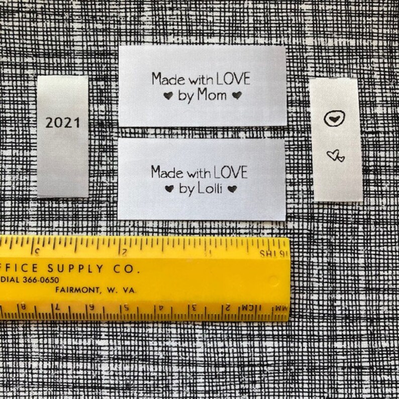 Puede incluir: Cuatro etiquetas de tela blanca con texto negro. Las etiquetas dicen "2021", "Made with LOVE ❤ by Mom ❤", "Made with LOVE ❤ by Lolli ❤" y un s&iacute;mbolo de coraz&oacute;n. Una regla amarilla es visible en la parte inferior de la imagen.