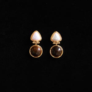 May include: A pair of gold-coloured earrings. Each earring has a round, dark brown stone set in a gold frame, with a white, triangular stone above. The earrings are shown against a black background.