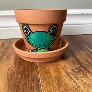 May include: A terracotta ceramic plant pot and saucer set. The pot features a hand-painted green frog face with black and white eyes. The frog design is centered on the front of the pot. The pot and saucer are on a wooden surface.