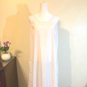May include: A white, sleeveless nightgown displayed on a mannequin. The nightgown has a simple, flowing design with a round neckline. The fabric appears lightweight and semi-sheer, suggesting a comfortable fit. The background is a light yellow wall.