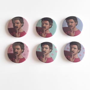 May include: Set of six round pinback buttons featuring a portrait of a man with dark hair and a red shirt, with his finger on his lips. The backgrounds vary in color, including pink, blue, and purple, some with heart patterns.