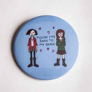 May include: A round, light blue button featuring cartoon illustrations of two girls with the text "You're the Jane to my Daria" and small pink hearts. One girl wears a red jacket, the other a green jacket.