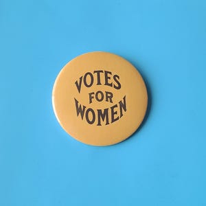 Puede incluir: Un botón redondo de color amarillo mostaza con las palabras "VOTES FOR WOMEN" en una fuente vintage estilizada. El botón está centrado sobre un fondo azul brillante, creando un contraste llamativo.