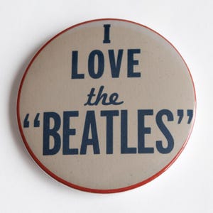 May include: A circular badge with a tan face and a red edge. The words "I LOVE the "BEATLES"" are printed in dark blue. A retro-style collectible item.