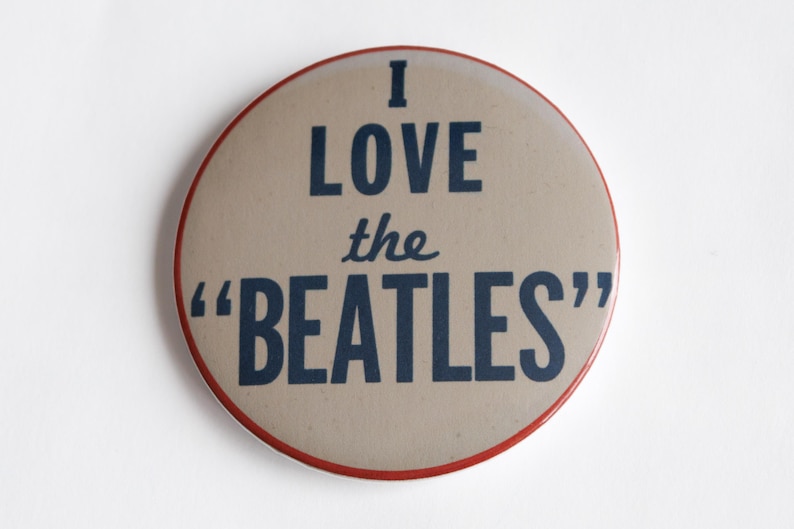 May include: A circular badge with a tan face and a red edge. The words "I LOVE the "BEATLES"" are printed in dark blue. A retro-style collectible item.