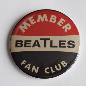 May include: A circular badge with a red upper section, a beige middle, and a black lower section. The text reads "MEMBER" in white, arched at the top, "BEATLES" in white across the middle, and "FAN CLUB" in white at the bottom.