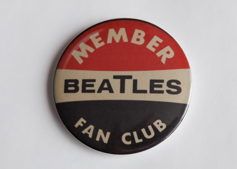 May include: A circular badge with a red upper section, a beige middle, and a black lower section. The text reads "MEMBER" in white, arched at the top, "BEATLES" in white across the middle, and "FAN CLUB" in white at the bottom.