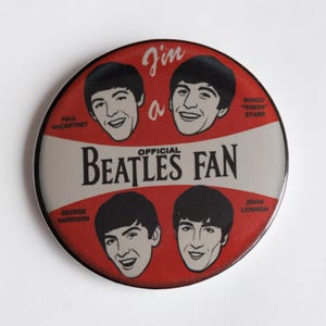 May include: A circular badge with a red and white design, featuring illustrations of The Beatles. The badge includes the text "I'm a Beatles Fan" and portraits of Paul McCartney, John Lennon, George Harrison, and Ringo Starr.