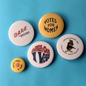 May include: Five circular badges with different slogans and graphics. One reads "D.A.R.E. to resist drugs and violence." Another says "Votes for Women." Other badges include "Watch Out Vote" and "Nuclear Power? No Thanks."