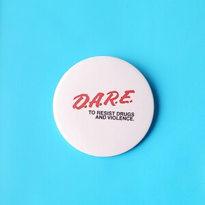May include: A white, circular button with the red text "D.A.R.E." above "TO RESIST DRUGS AND VIOLENCE." The button is set against a bright blue background. The button is a classic design.