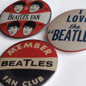 May include: Three vintage Beatles fan club badges. One badge reads "I LOVE the 'BEATLES'" in navy blue on a tan background. Another badge says "BEATLES FAN" with band member portraits. The third badge says "MEMBER BEATLES FAN CLUB".