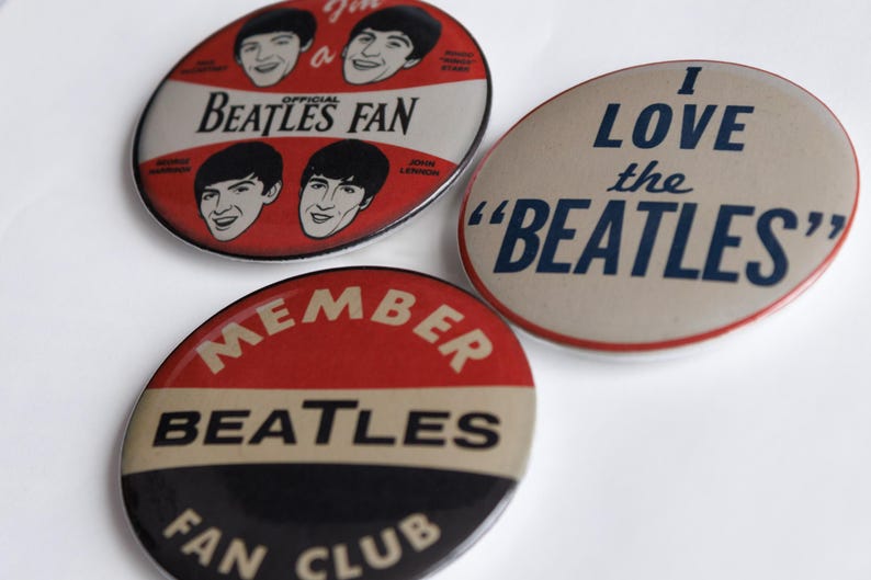 May include: Three vintage Beatles fan club badges. One badge reads "I LOVE the 'BEATLES'" in navy blue on a tan background. Another badge says "BEATLES FAN" with band member portraits. The third badge says "MEMBER BEATLES FAN CLUB".