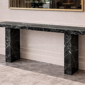 May include: A black marble console table with white veining. The rectangular table has a long top supported by two thick, rectangular legs. The table is set against a neutral wall with a gold-framed mirror above.