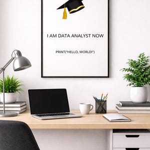 May include: A framed print with a black border displays a graduation cap illustration and the text "I AM DATA ANALYST NOW" and "PRINT("HELLO, WORLD!")". A laptop, desk lamp, and potted plants are on a wooden desk.