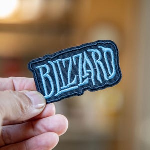 Puede incluir: Un parche rectangular con la palabra "Blizzard" bordada en azul claro sobre un fondo azul oscuro. El parche tiene un borde azul oscuro y está sostenido en una mano.