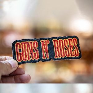 Könnte beinhalten: Gestickter Aufnäher mit dem Text "GUNS N' ROSES" in roter und gelber Schrift auf schwarzem Hintergrund. Der Aufnäher hat einen schwarzen Rand und wird von einer Hand gehalten. Zum Aufnähen auf Kleidung oder Taschen.