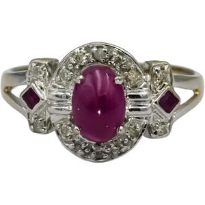 May include: A vintage ring with a large oval ruby gemstone, encircled by small diamonds set in silver-tone metal. The ring features two smaller ruby accents on the sides, creating a classic and elegant design.