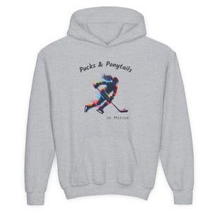 Puede incluir: Una sudadera con capucha gris claro con el texto "Pucks & Ponytails" y una ilustraci&oacute;n de un jugador de hockey en movimiento. La ilustraci&oacute;n presenta un dise&ntilde;o colorido y estilizado de una persona con una cola de caballo jugando al hockey.