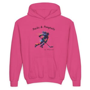 Puede incluir: Una sudadera con capucha rosa intenso con un bolsillo delantero y una capucha. La sudadera presenta el texto "Pucks & Ponytails" sobre una colorida ilustraci&oacute;n de una jugadora de hockey en movimiento. Debajo de la ilustraci&oacute;n est&aacute; el texto "In Motion."