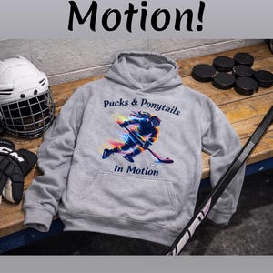 Puede incluir: Una sudadera con capucha gris o rosa con el texto "Pucks & Ponytails In Motion!" y un gr&aacute;fico de jugador de hockey. La imagen muestra a una persona joven usando la sudadera, con equipo de hockey en el fondo.