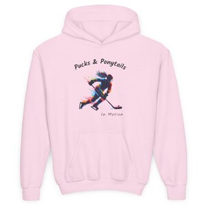 Puede incluir: Sudadera con capucha rosa claro con bolsillo delantero y capucha. El gr&aacute;fico frontal presenta a una jugadora de hockey en movimiento con el texto "Pucks & Ponytails In Motion". El dise&ntilde;o utiliza un efecto colorido y estilizado.