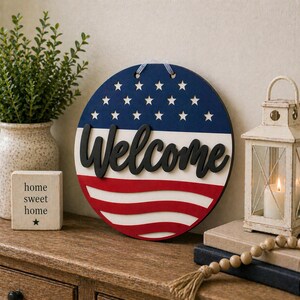 Puede incluir: Un letrero redondo de madera con un dise&ntilde;o patri&oacute;tico. La parte superior es azul con estrellas blancas, el medio es blanco y la parte inferior tiene rayas rojas. La palabra "Welcome" est&aacute; en escritura negra. Un elemento decorativo para el hogar.