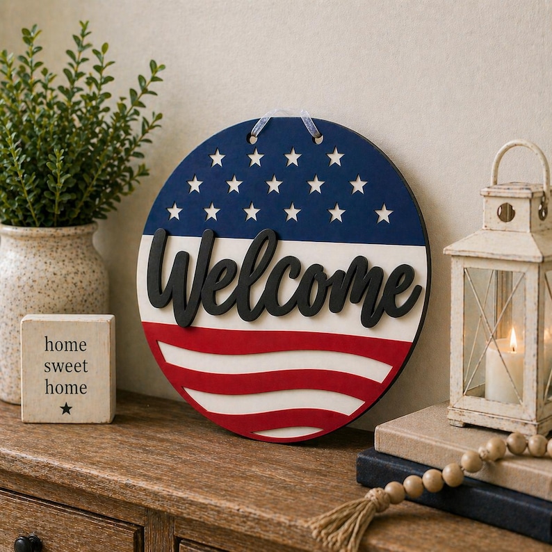 Puede incluir: Un letrero redondo de madera con un dise&ntilde;o patri&oacute;tico. La parte superior es azul con estrellas blancas, el medio es blanco y la parte inferior tiene rayas rojas. La palabra "Welcome" est&aacute; en escritura negra. Un elemento decorativo para el hogar.