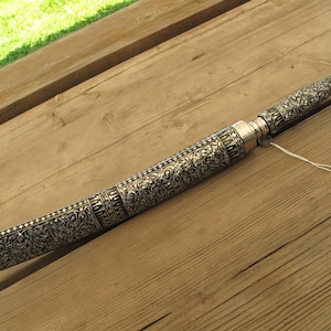 May include: An ornate silver-colored object with intricate floral patterns. The object is long and cylindrical, possibly a container or decorative item. It rests on a wooden surface, suggesting a display or storage piece.